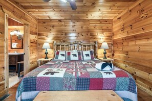 Ein Schlafzimmer in einer Holzhütte mit einem Bett, zwei Nachttischlampen, einem Holz-Kopfteil und einer Patchworkdecke mit Tiermotiven.