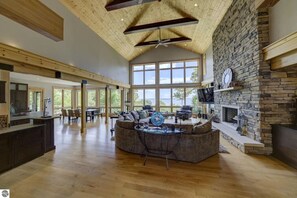 A spacious living area with a stone fireplace, large windows, and a high ceiling with exposed beams. - Spyglass Ridge Retreat – 6BR Luxury Bay View Pines Estate in Suttons Bay (Suttons Bay)