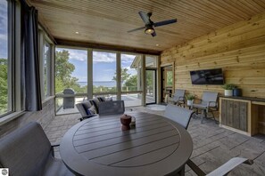 A modern outdoor dining area with a round table, chairs, a ceiling fan, and a TV mounted on the wall. - Spyglass Ridge Retreat – 6BR Luxury Bay View Pines Estate in Suttons Bay (Suttons Bay)