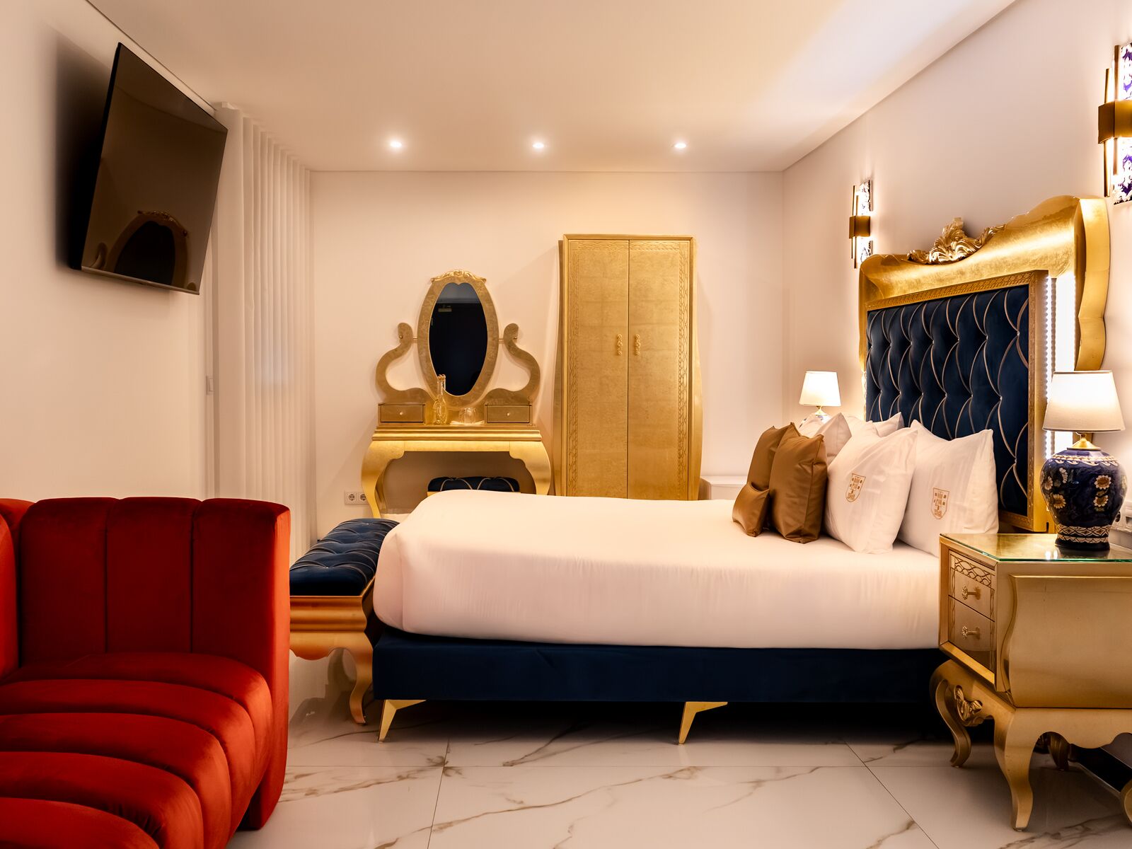 Habitació d'hotel amb un llit, una cadira vermella, un tocador daurat i una televisió penjada a la paret.