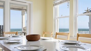 A dining table set for four with white plates, silverware, and a woven bowl. The table is positioned near large windows with ocean views. - aOceanfront | Incredible Views | Steps to the Beach (Surf City)