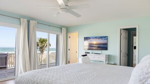A bedroom with a large bed, a ceiling fan, a painting of a sunset, a door, and a view of the ocean. - aOceanfront | Incredible Views | Steps to the Beach (Surf City)