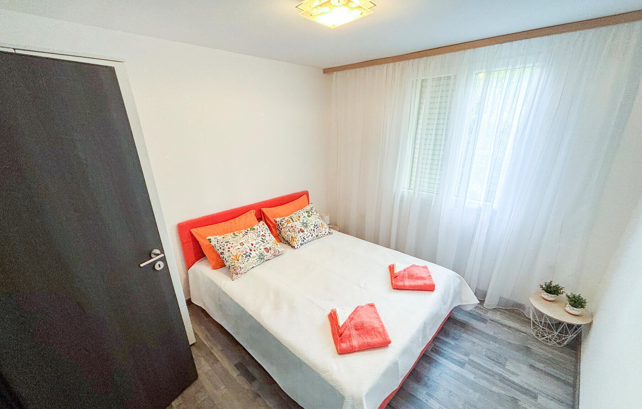 A bedroom with a bed, curtains, a small table with plants, and a door.