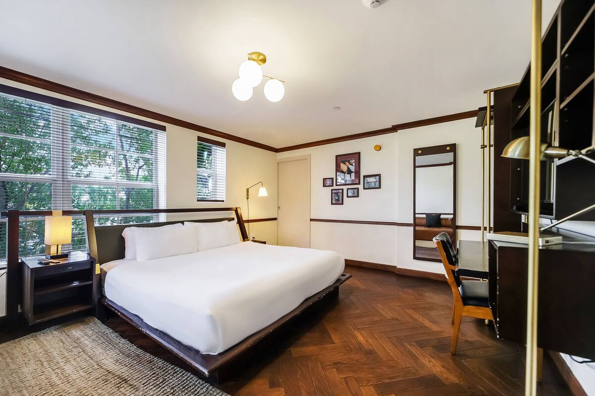 A hotel room with a large bed, a desk, a chair, a desk lamp, a mirror, and a window with blinds.