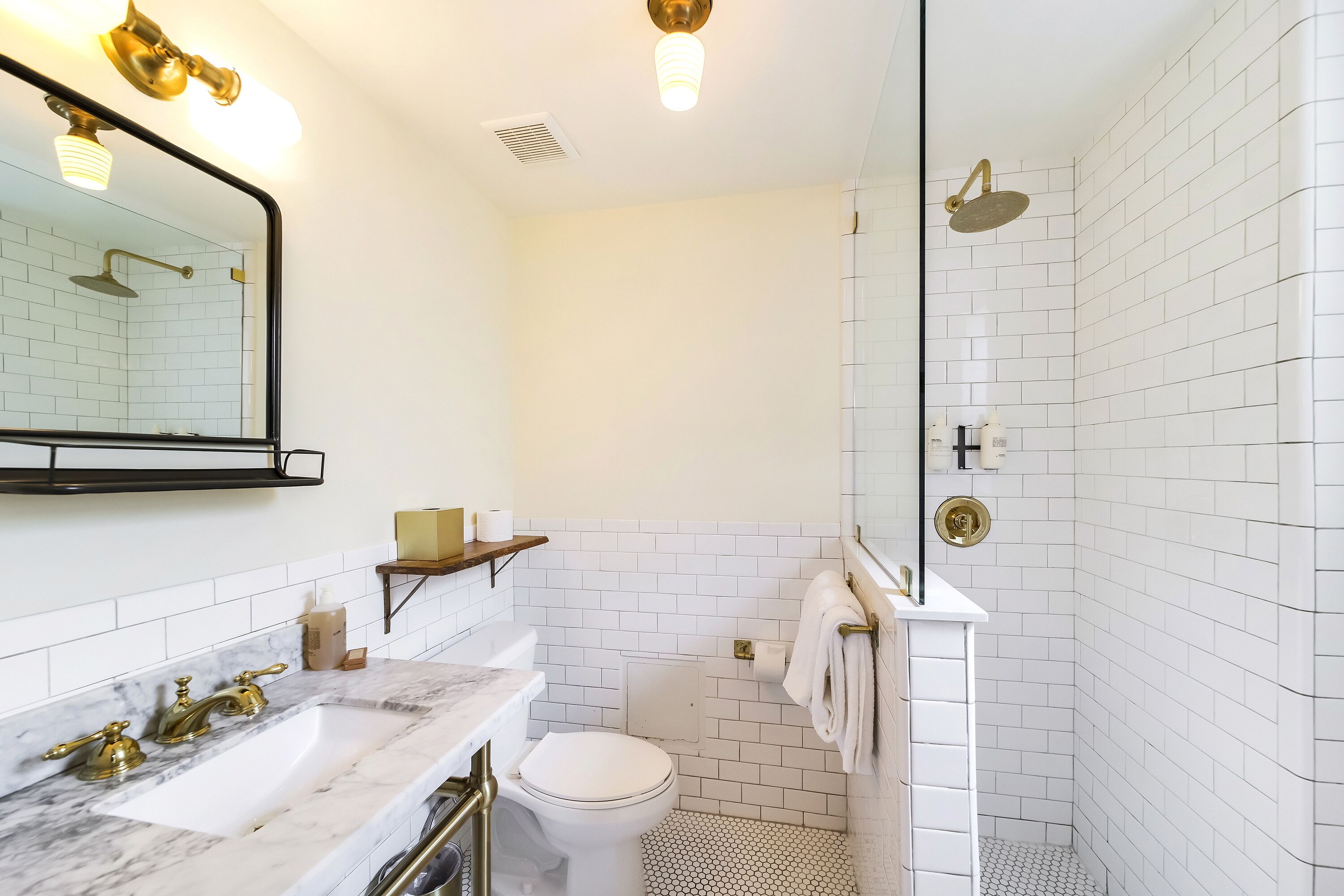 A bathroom with a white tiled shower, a toilet, a sink with gold fixtures, and a mirror with a black frame.
