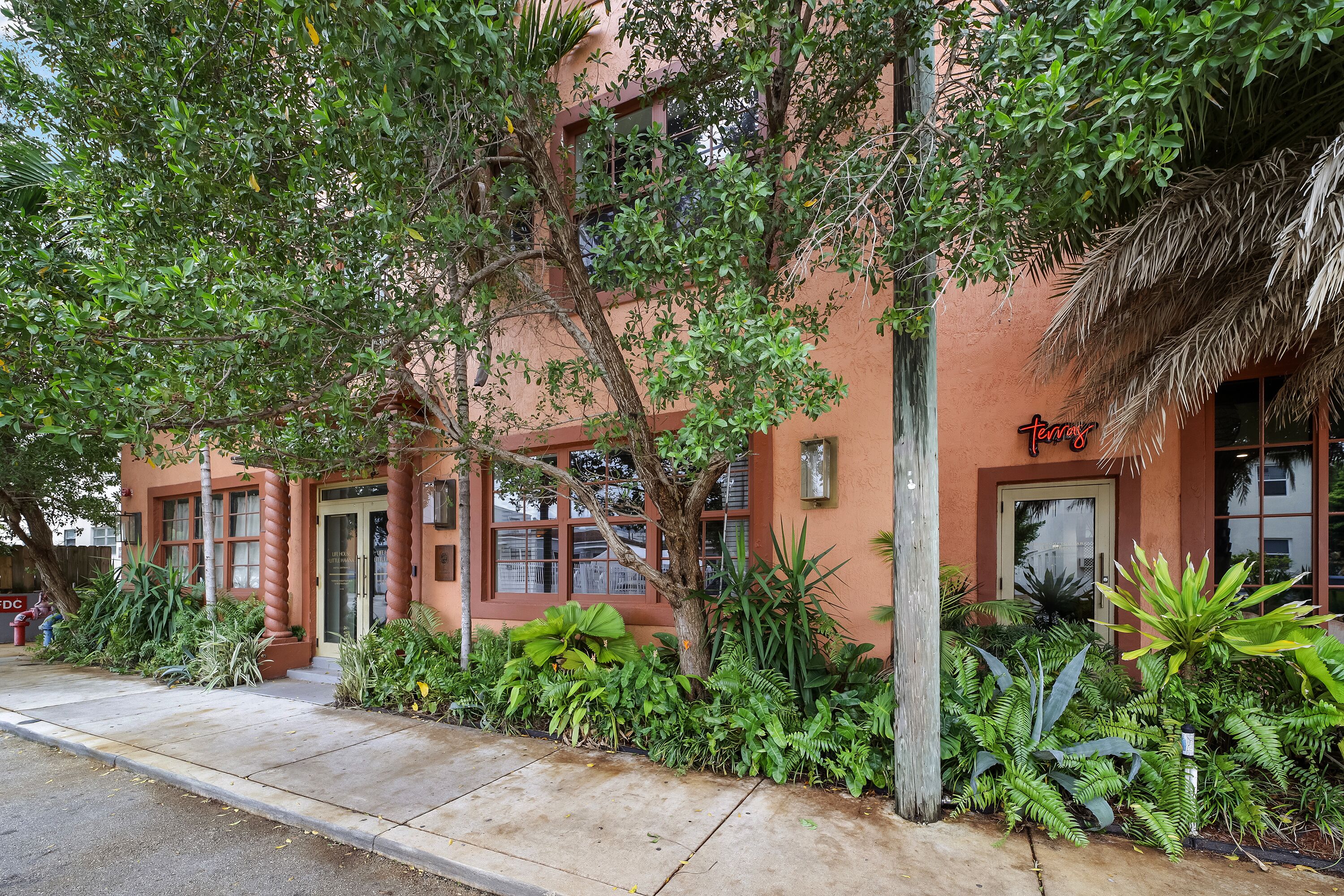 A building with a red facade, a sign that reads 'terraces,' and a variety of tropical plants around the entrance.
