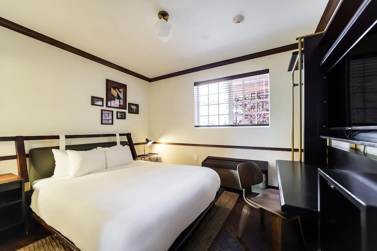 A hotel room with a bed, desk, chair, window, and artwork on the wall.