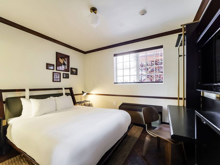 A hotel room with a bed, desk, chair, window, and artwork on the wall.