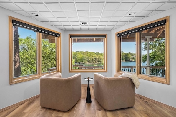 A room with large windows, two beige chairs, a small table, and a view of a lake and trees.