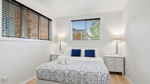 Un dormitorio con cama, dos mesitas de noche, una ventana con persianas y vistas al exterior.
