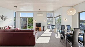 Ein modernes Wohnzimmer mit Sofa, Fernseher, Esstisch, Stühlen und großen Fenstern.