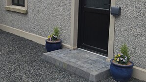 Un edificio con fachada texturizada, una puerta negra, un ventanal pequeño y dos macetas con plantas.