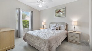Ein Schlafzimmer mit einem Bett, zwei Nachttischen mit Lampen, einem Deckenventilator, einem Fenster mit Vorhängen und einem gerahmten Bild an der Wand.