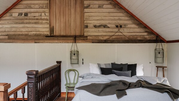 A bedroom with a sloped ceiling, a wooden bed frame, a green chair, and two hanging lanterns. - Demi Barn - Unique Historic Country Barn (Daylesford)