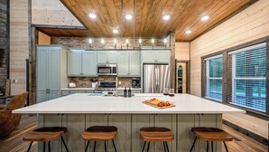 A modern kitchen with a large island, bar stools, and a brick wall. - Retreat w/ Sauna, Hot Tub, Games & Fenced Dog Yard! (Broken Bow)