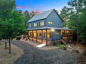 A two-story house with a porch, a staircase leading to the upper level, and a gravel driveway. - Retreat w/ Sauna, Hot Tub, Games & Fenced Dog Yard! (Broken Bow)