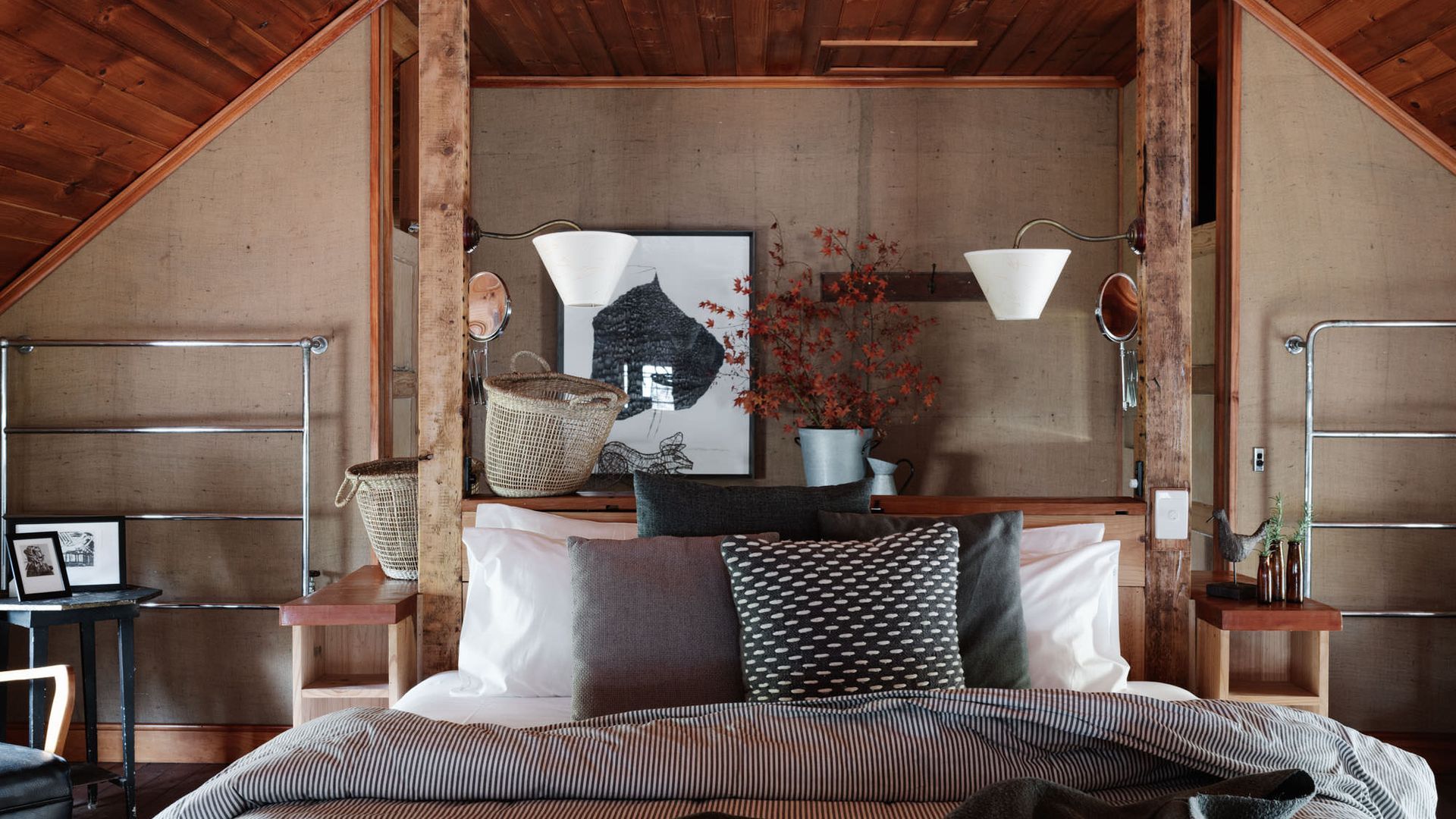 A cozy bedroom with a sloped ceiling, a bed with striped bedding, a wooden nightstand, a wall-mounted lamp, and a framed artwork.