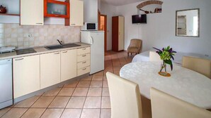 Een compacte keuken met een spoelbak, magnetron en kastjes. Er is een eethoek met een tafel en stoelen, en een televisie bevestigd aan de muur.