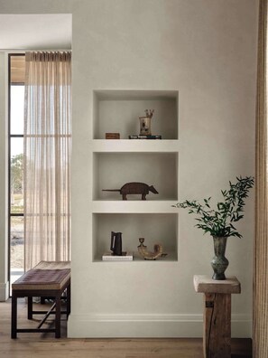 Una habitació amb una banca de fusta, una estanteria amb llibres i objectes decoratius, i una finestra amb cortines de tela fina.