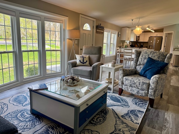 A living room with a glass coffee table, a grey armchair, a patterned armchair, and a view of the outdoors through large windows.
