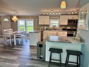 A kitchen with a dining area, a chandelier, a sign reading 'BEACH HOUSE,' and a view of the outdoors.