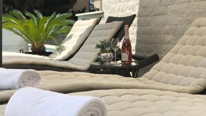 Une chaise longue, une serviette enroulée, une table en verre avec une bouteille et des verres, et une plante en pot.