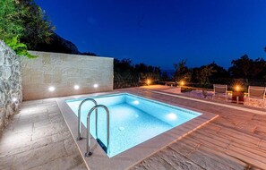A rectangular swimming pool with metal railings, surrounded by a stone wall and wooden deck.