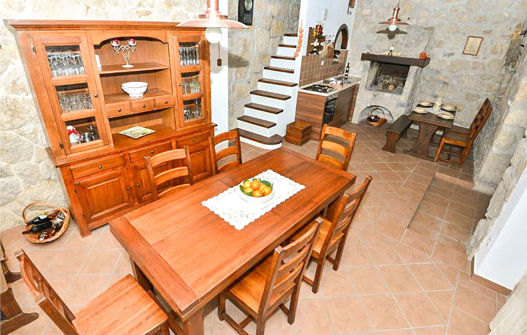 A dining area with a wooden table, chairs, a cabinet with glass doors, a staircase, and a stone wall.