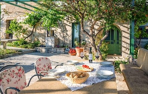 An outdoor dining area with stone walls, a green door, and a table set with plates, a basket of bread, and a bottle of wine.