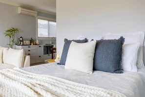 Et soveværelse med en seng, puder, et natbord, en lampe, en plante og et køkken i baggrunden.