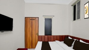 Une chambre d’hôtel avec un lit, une porte en bois, une petite fenêtre, un téléviseur fixé au mur et un banc rouge.