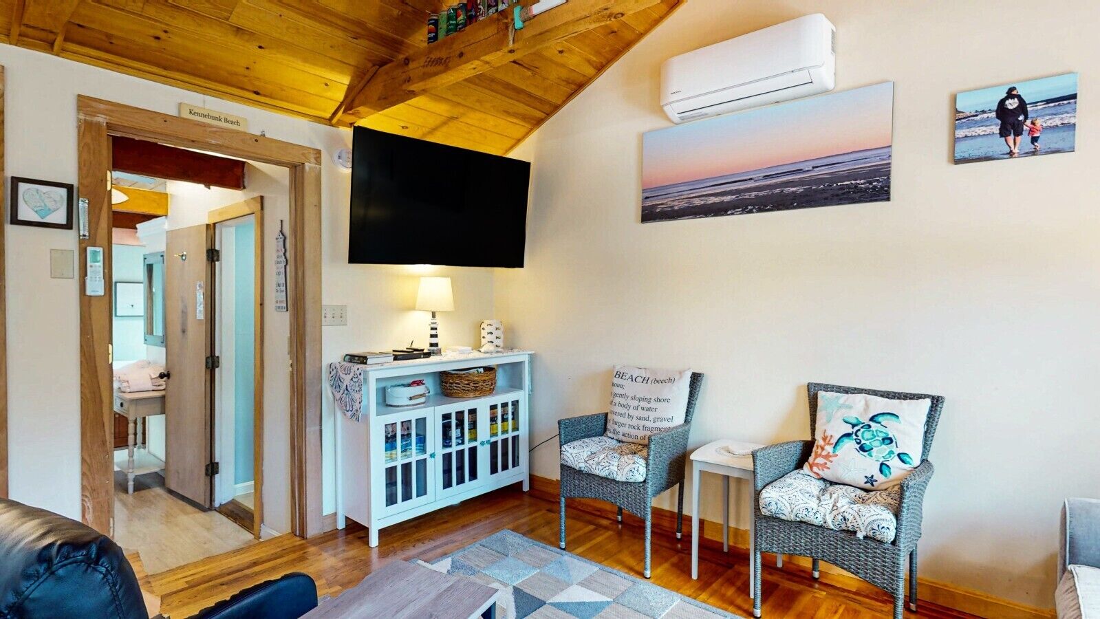 Een gezellige woonkamer met een houten plafond, een flatscreen televisie, twee rieten fauteuils, een klein tafeltje en een wandairconditioner.