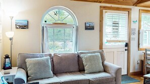 A cozy room with a sofa, a lamp, a window with a view, and a door. - Coastal Shorelands Retreat + Walk-to-Beach Access and Pool (Kennebunk)