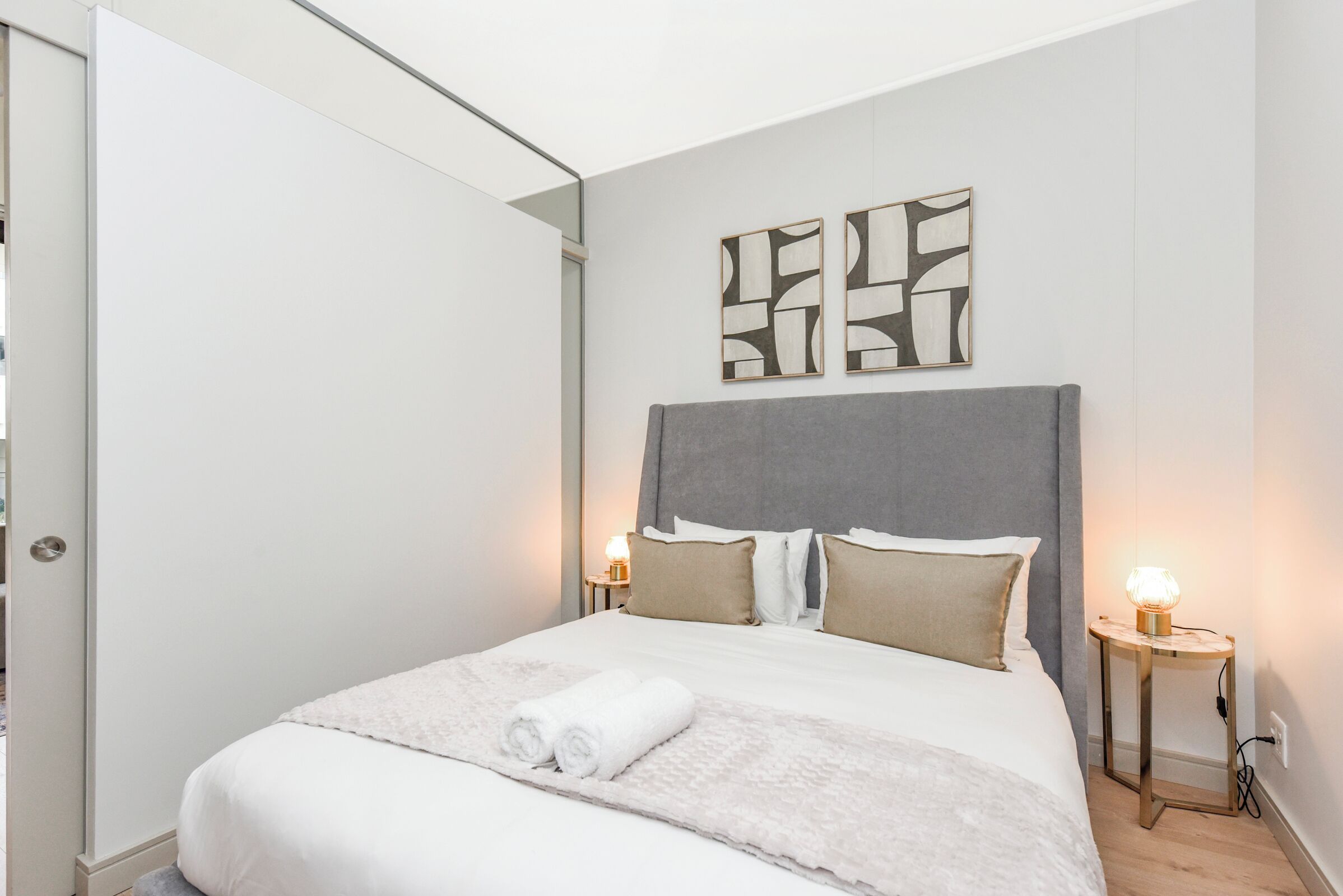 A neatly made bed with a grey headboard, two bedside tables with lamps, and abstract wall art.