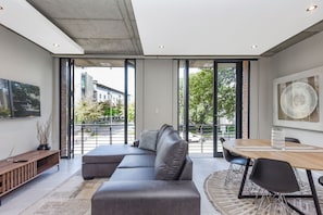 Ein modernes Wohnzimmer mit einer Eckcouch, einem Esstisch mit Stühlen und großen Fenstern mit Blick auf Gebäude und Bäume.