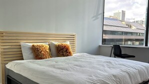 Een moderne slaapkamer met een groot bed, een houten hoofdbord en uitzicht op gebouwen door een raam.