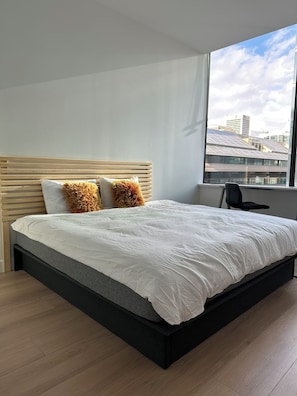 A modern bedroom with a large bed, a wooden headboard, and a view of buildings through a window. - Curated Comfort: 1BR Loft | King+Double Beds (Vancouver)