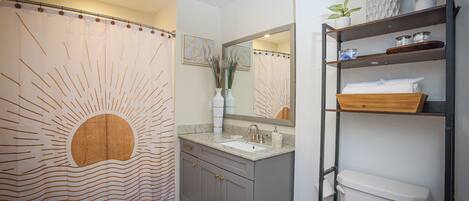A bathroom with a shower curtain, sink, mirror, toilet, and shelves with toiletries.