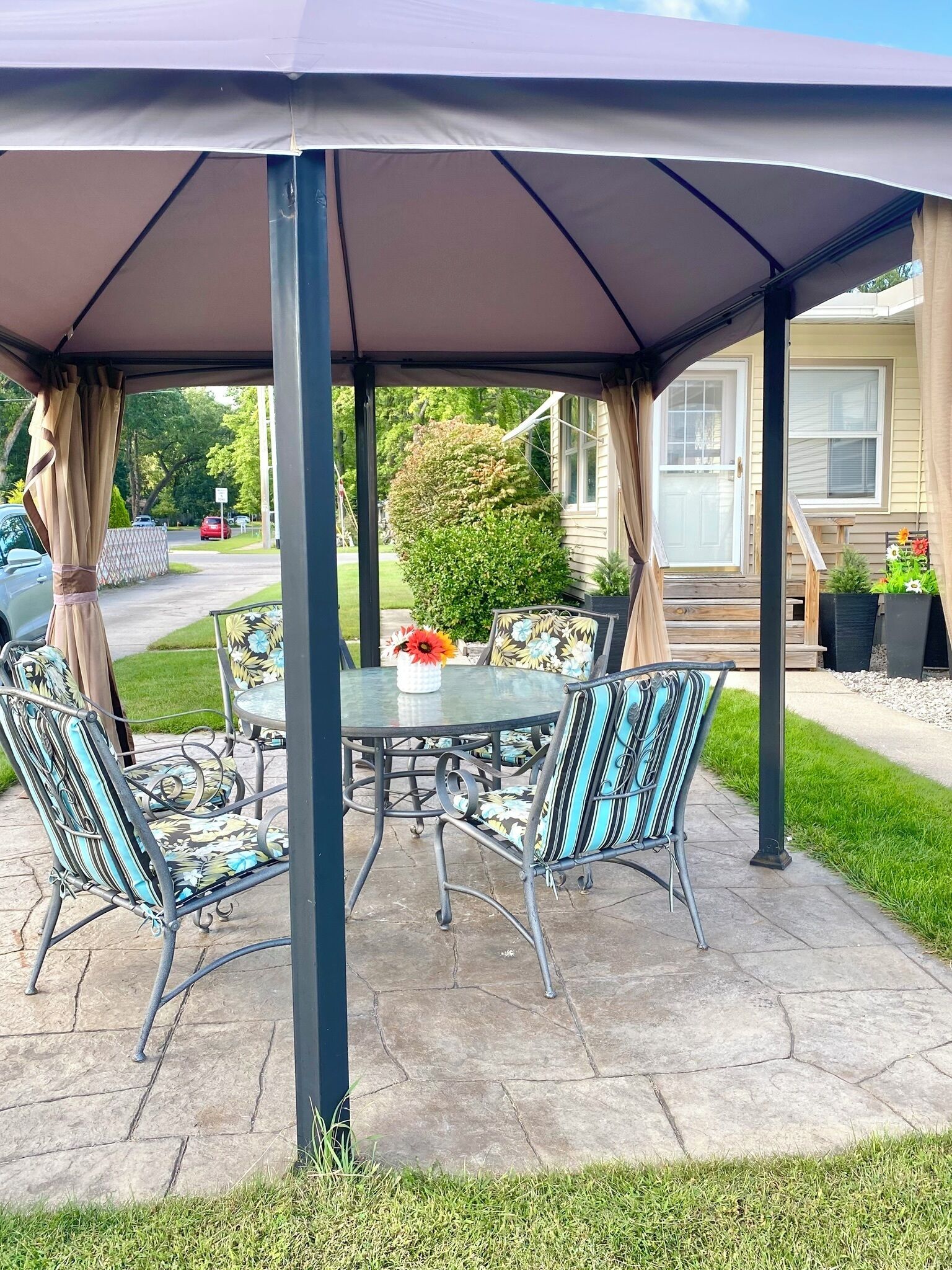 A covered patio area with a table and chairs, a flower vase, and a view of a residential street.