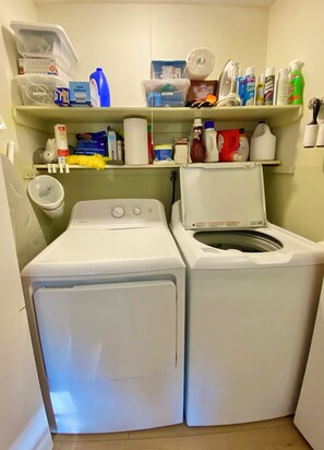 A laundry room with two front-loading washing machines, shelves above containing cleaning supplies, and a toilet paper holder on the wall. - Under The Cherry Boon: A Traverse City Dream (Traverse City)