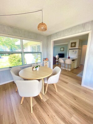 A dining area with a round table and four chairs, a hanging light fixture, and a view of a kitchen and outdoor area through an open doorway. - Under The Cherry Boon: A Traverse City Dream (Traverse City)