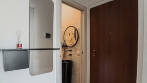 Un couloir avec un mur blanc, un plancher en bois, une porte, un miroir, une poubelle et une petite étagère sur laquelle se trouve une bouteille.