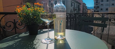Una mesa con una botella de vino y una copa, rodeada de flores en macetas, bajo un cielo despejado.
