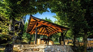A wooden gazebo with a stone base, surrounded by trees and a clear sky.