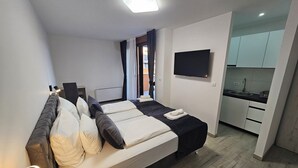 A modern hotel room with a bed, bedside table, lamp, TV, and a kitchenette.