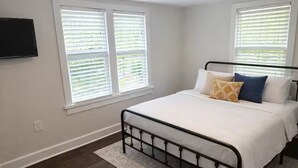 A bedroom with a bed, two windows with blinds, a television on the wall, and a rug. - The Downtown Cedar - walk to Government St & Washington (Ocean Springs)