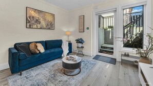 Uma sala de estar com um sofá azul, uma mesa de centro redonda, um quadro na parede e uma escada que dá para o exterior.