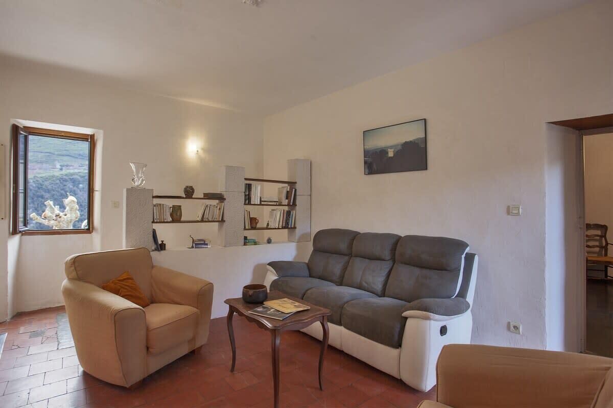 A living room with a sofa, armchair, and a small table. There is a bookshelf with books and decorative items. A window with a view of a landscape is visible.