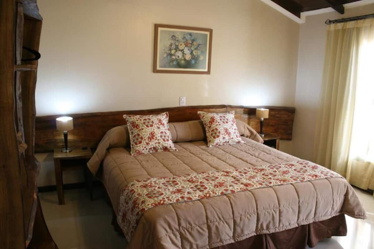 A bedroom with a bed, two bedside tables, a lamp, a painting on the wall, and a window with curtains.