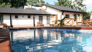 A swimming pool with a tiled edge, wooden lounge chairs, and a building with a tiled roof and windows. - Posada Del Jacarandá (Puerto Iguazu)